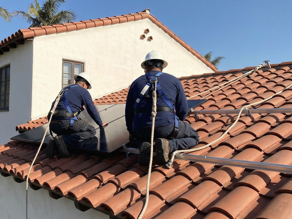 Two installers in harnesses roped to a roof anchor, kneeling on a red tile roof as they set a black solar panel on its rails