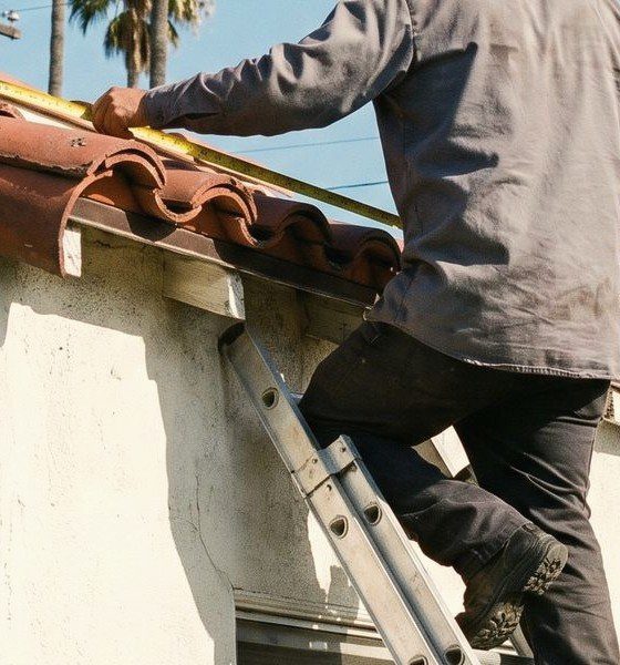 A worker on a ladder running a tape measure along the edge of a red clay tile roof